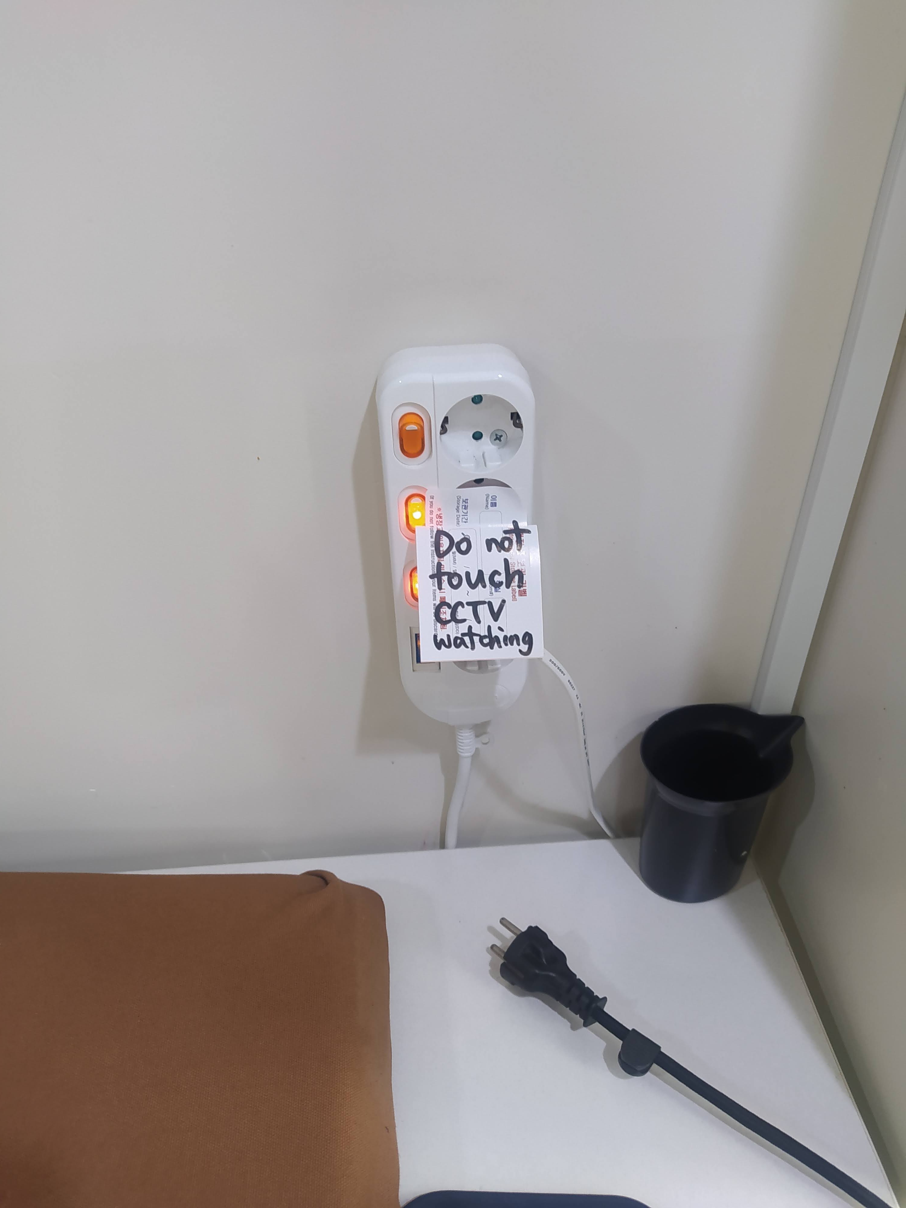 Figure 4.4. «&nbsp;Ne pas toucher, la caméra de surveillance veille&nbsp;» (notre trad.). Photographie d’une multiprise murale avec une étiquette grossière priant en anglais de ne pas y toucher. Autrice&nbsp;: Assia Musitelli Chabane, 2023